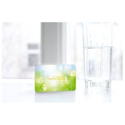 A glass of water placed on the Raumvital "Seelenquell" Vitalcard, with a subtle glow, suggesting spiritual clarity.